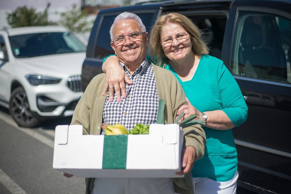 A tan man with white hair and an older woman with blond hair smile while holding a box of produce. They can enjoy their retirement and social security in Las Vegas.