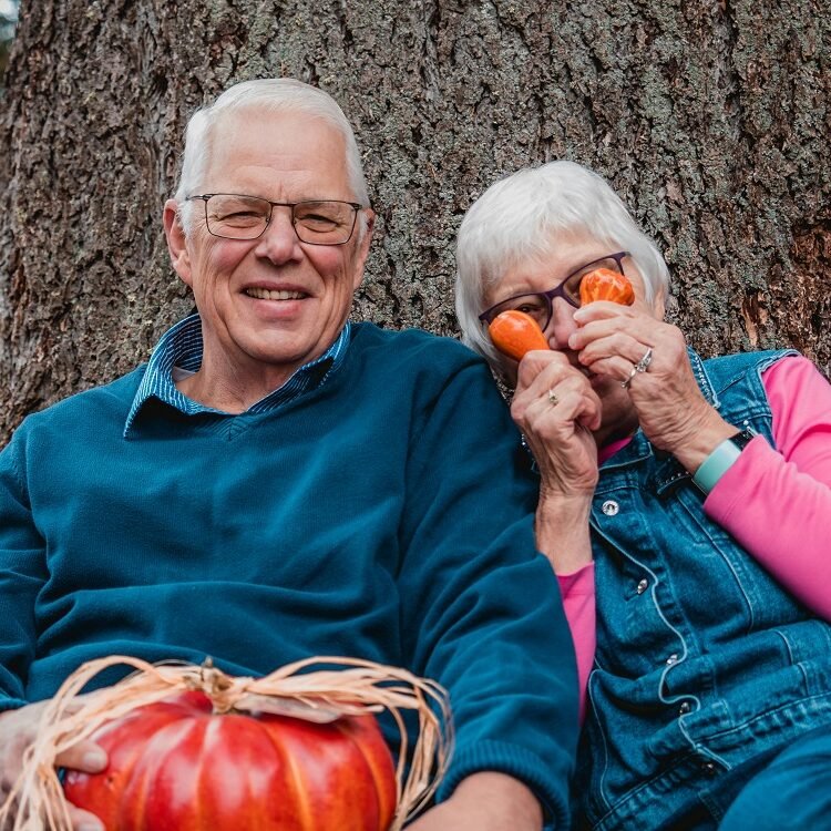 An older white man and woman with white hair sitting in front of a tree. The woman holds pumkins over her eyes.